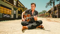 Musician seated with guitar in a rustic Western-themed town, Wimberley, TX